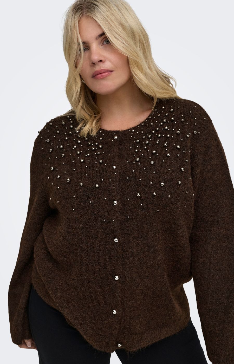 A woman wearing the Only Carmakoma Miggie Cardigan in Coffee Bean, a brown fuzzy knit cardigan sweater with silver rhinestone embellishments across the chest and front buttons, paired with black pants, against a light gray studio background.
