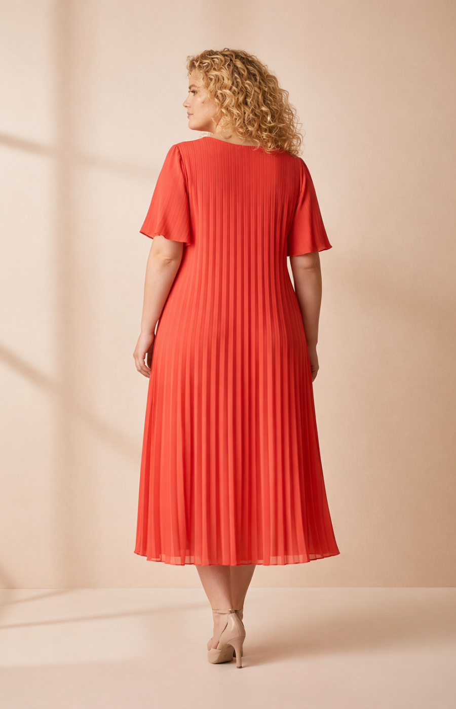 A full-length front view of a woman with shoulder-length curly blonde hair wearing a coral pleated midi dress with short flutter sleeves. She stands in high heels against a soft beige studio background with warm, even lighting.