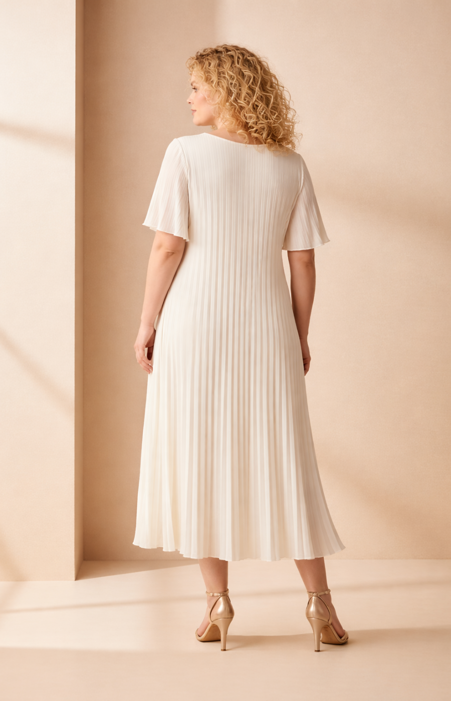 A full-length rear view of a woman with shoulder-length curly blonde hair wearing a cream pleated midi dress with short flutter sleeves. She stands in high heels against a soft beige studio background with warm, even lighting.