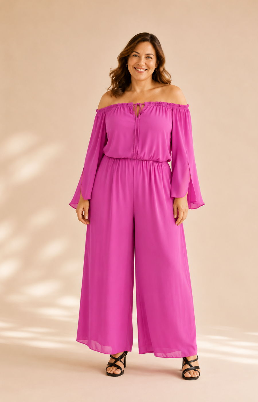 A woman modeling a bright magenta off-the-shoulder jumpsuit with long sleeves and wide legs, standing against a warm beige studio backdrop with soft shadow overlays.