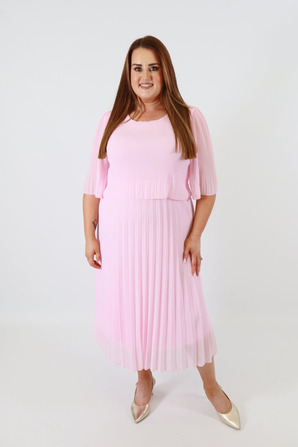 Plus size woman wearing a pink pleated dress with gold shoes on a white background