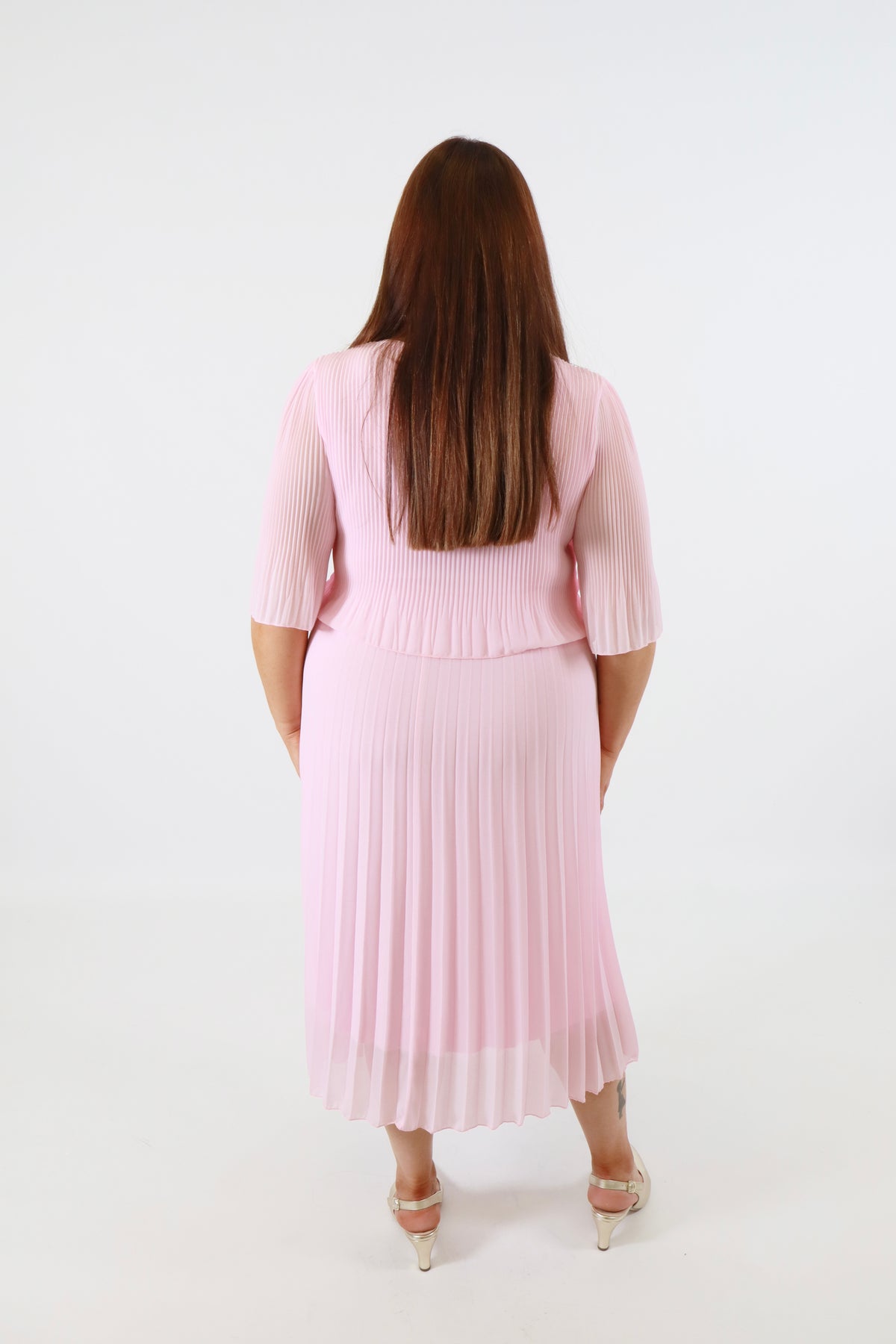 Plus size woman wearing a pink pleated dress with gold shoes on a white background