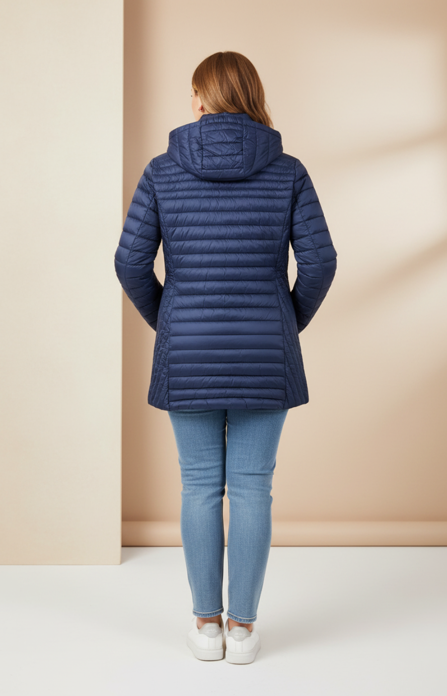 Plus size woman wearing a navy coat, light coloured jeans and white trainers on a cream background