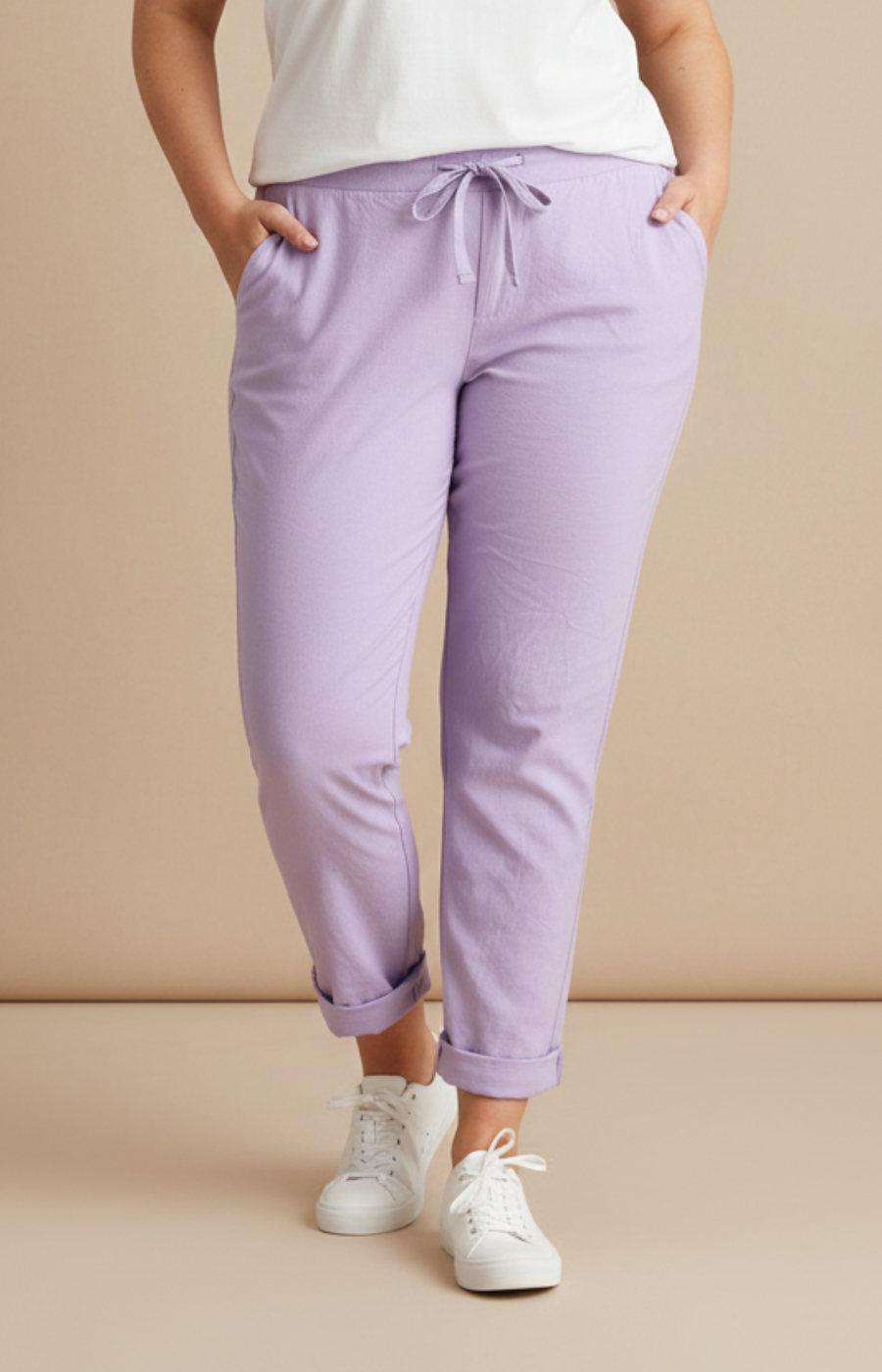 A plus size woman wearing lilac joggers and a white t-shirt on a cream background
