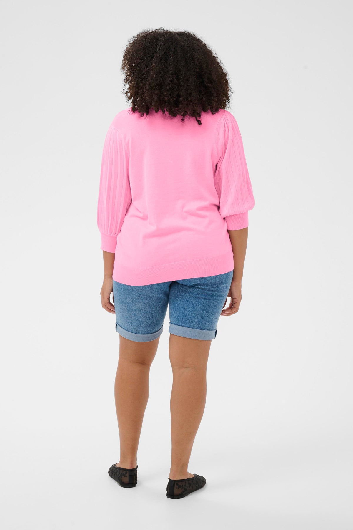 Person wearing a pink 3/4 length sleeve top and blue shorts on a white background