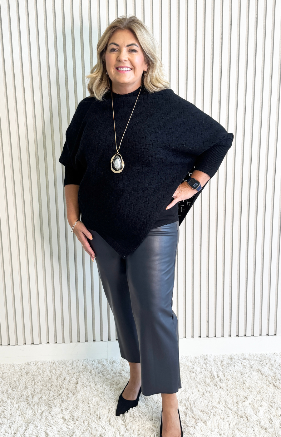 Woman wearing a black poncho and gray pants against a white paneled wall.