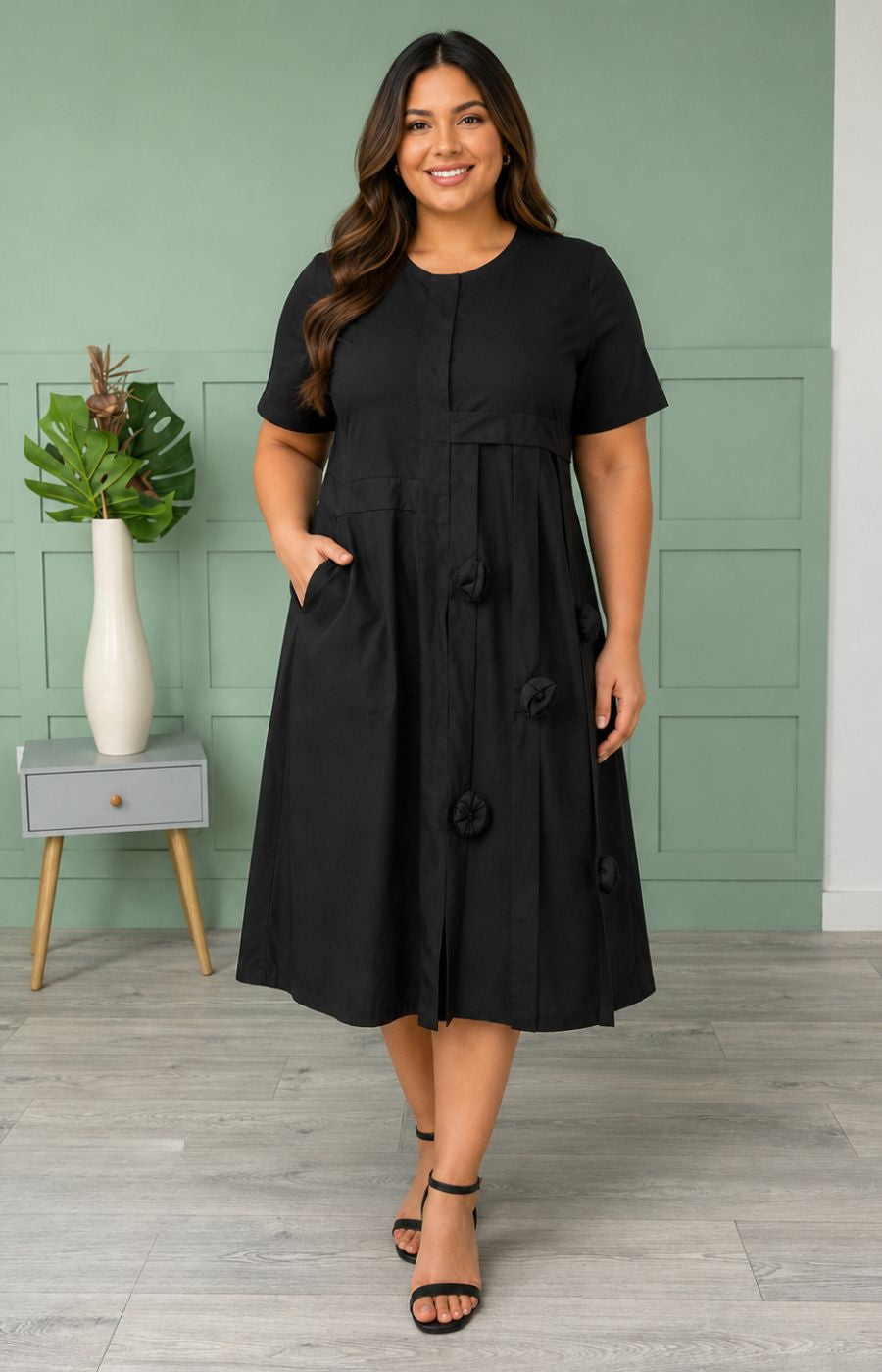 Woman wearing the Leah Dress in Black, a black dress standing in a room with a green wall and white vase.