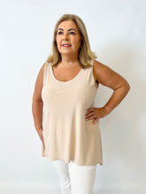Woman wearing a beige sleeveless top and white pants against a plain background