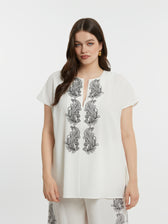 Woman wearing a white top with black floral embroidery with short sleeves and a round neckline with a V slit on a plain background