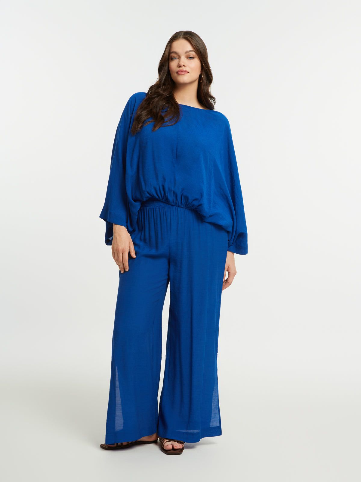 Woman wearing the Mat Slit Trouser in Blue, a high rise trouser with a wide leg and side slits, worn with a blue blouse on a white background