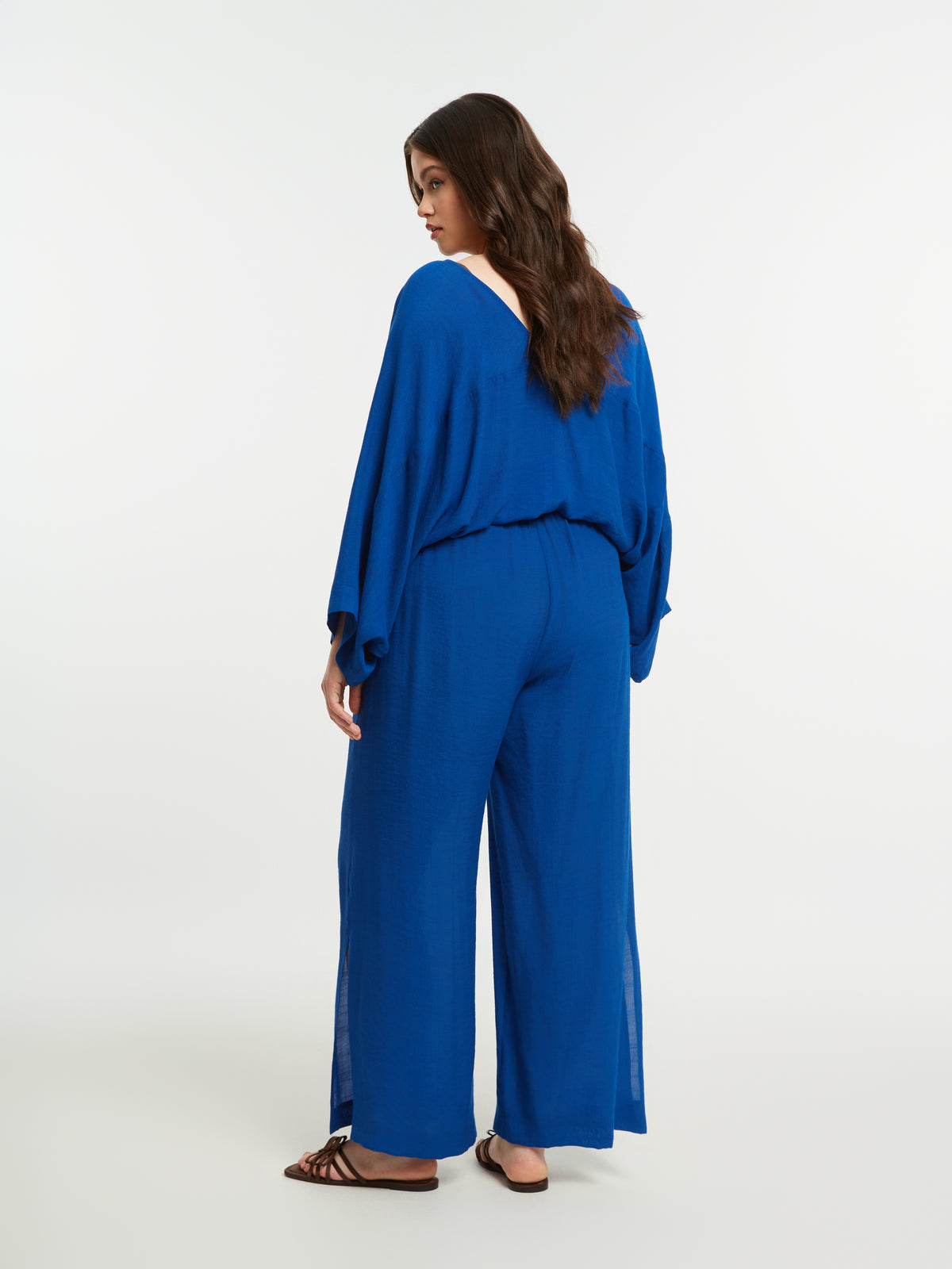 Woman wearing the Mat Slit Trouser in Blue, a blue, wide leg trouser with a pull up style and side slits, worn with a blue blouse on a white background