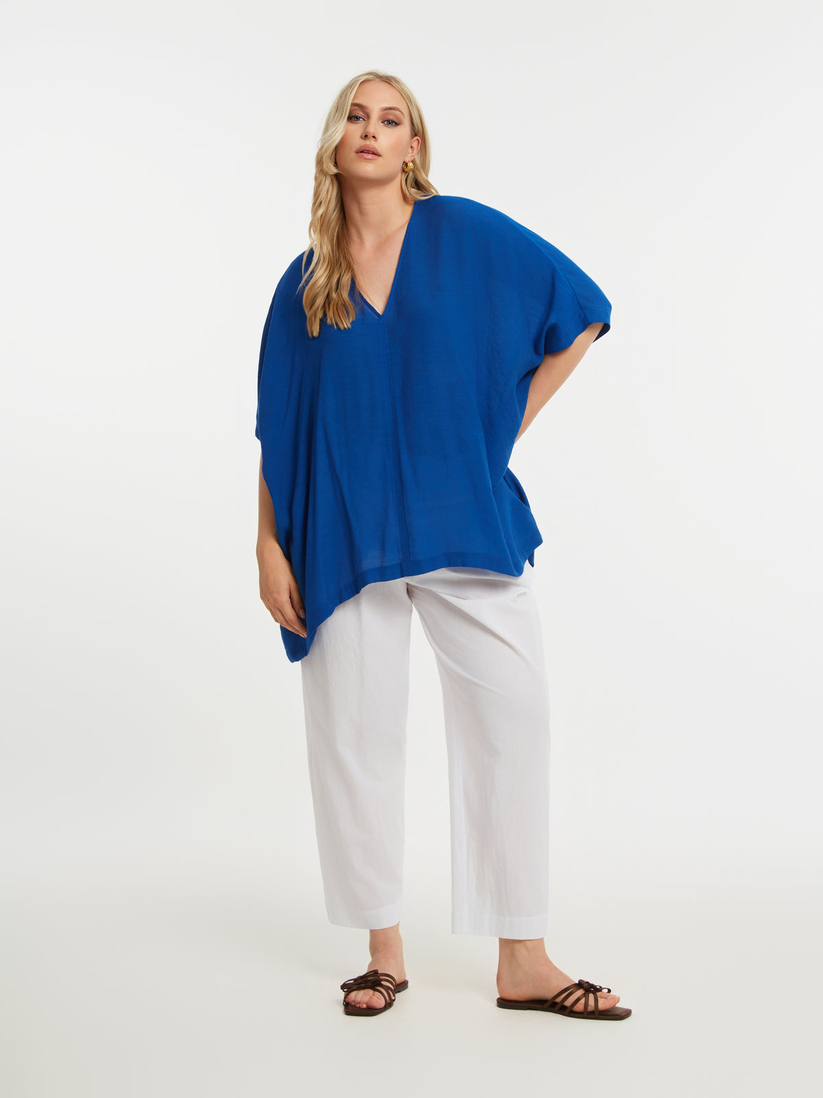 Woman wearing the Mat V Neck Blouse in Blue, a blue blouse with short sleeves, V neckline and a curved hem, and white trousers on a white background