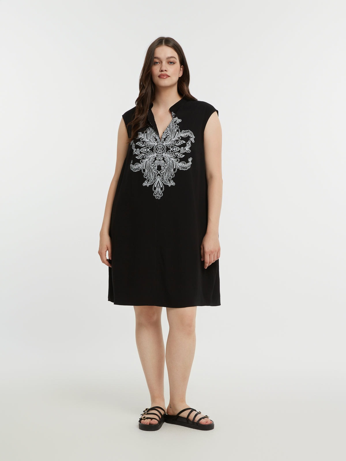 Woman wearing a black sleeveless dress with a V neckline and white embroidery on a white background