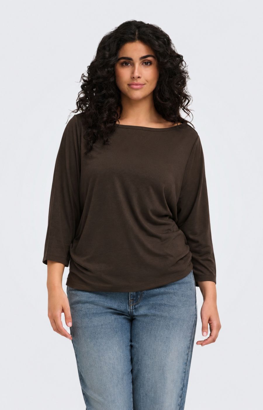 A woman with long, curly dark hair wearing the Only Carmakoma Carlinne Top in Coffee Bean, a dark brown boat-neck top and blue jeans, standing against a light gray studio background.