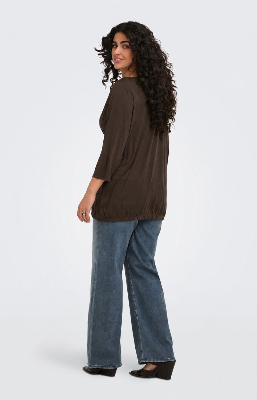 A woman with long curly dark hair is shown from a side-back angle wearing the Only Carmakoma Carlinne Top in Coffee Bean, a dark brown, loose-fitting three-quarter sleeve top and wide-leg blue jeans against a light gray studio background.