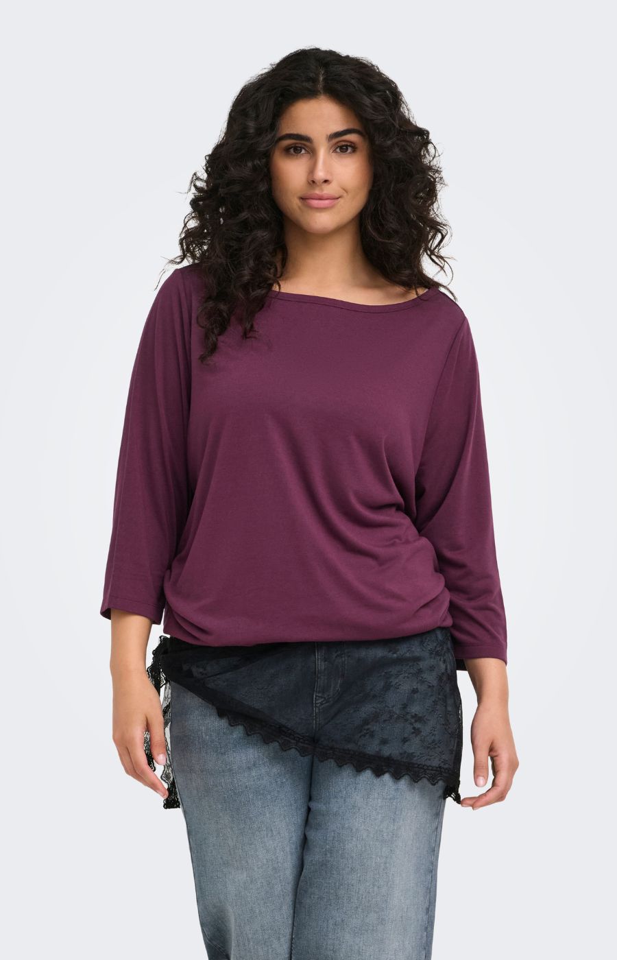 A woman modeling the Only Carmakoma Carlinne Top in Fig, a loose-fitting, three-quarter sleeve top in a deep plum purple color, paired with light-wash blue jeans and a black lace-trimmed skirt, against a plain light gray background.