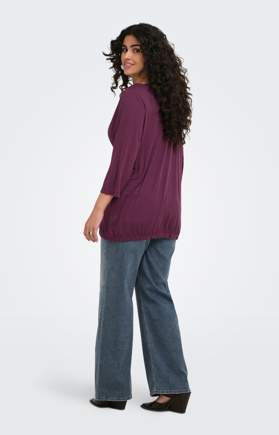 A woman wearing the Only Carmakoma Carlinne Top in Fig, shown from a side-back angle wearing a loose-fitting, three-quarter sleeve purple top and wide-leg blue jeans against a plain light gray studio background.