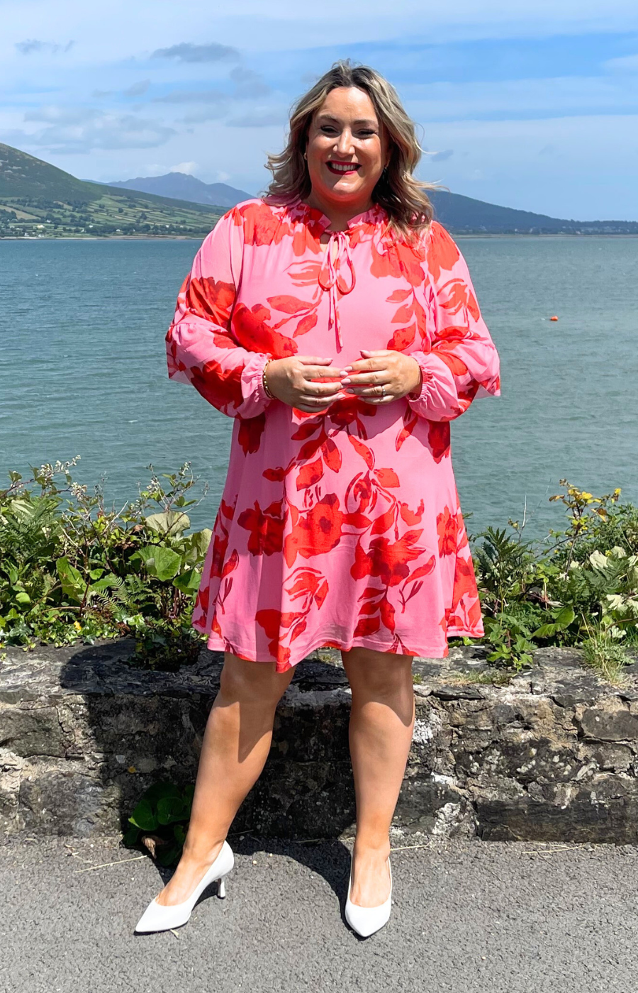 Woman in a pink floral dress standing by a scenic waterfront with mountains in the background.
