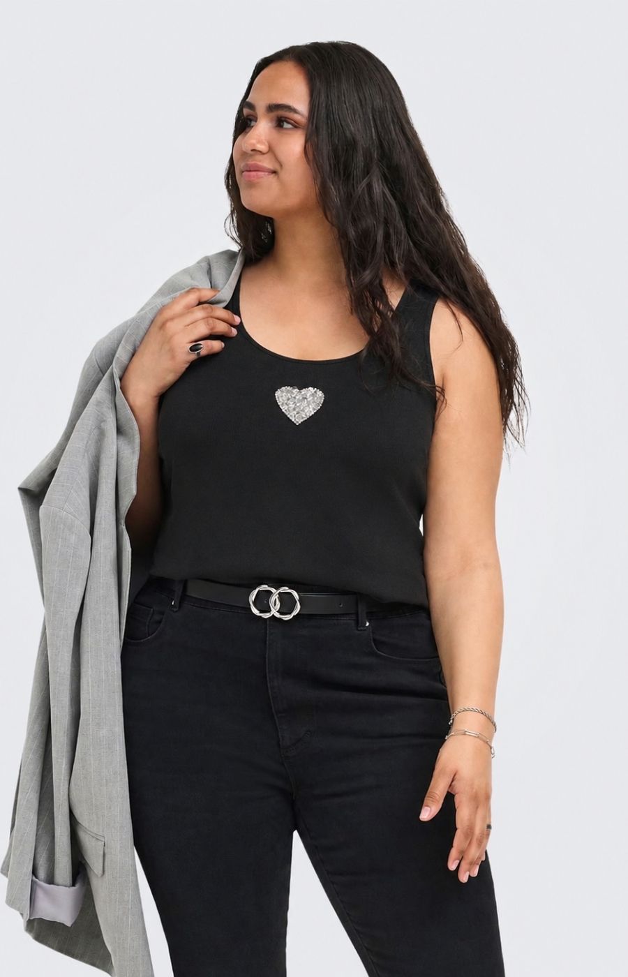 A woman modeling the Only Carmakoma Iris Vest Top in Black, a black sleeveless tank top with a sparkling silver heart emblem on the chest, paired with black jeans and a gray blazer draped over her shoulder, against a light gray background. This image was created using AI.