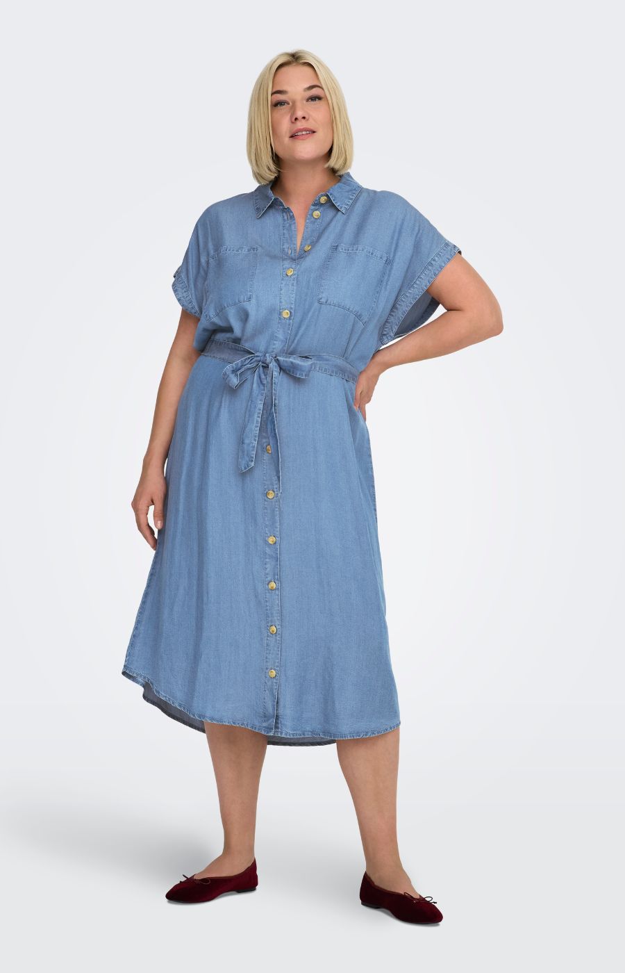 Woman wearing the Only Carmakoma Pema Denim Dress, a blue denim dress with a collared neckline, button down front, midi length and short sleeves on a white background