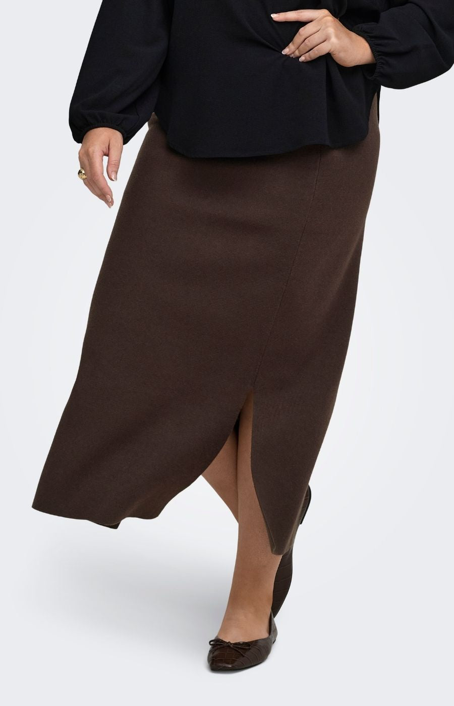 A woman wearing the Only Carmakoma Ribi Skirt in Brown, a dark brown maxi skirt with a front slit, paired with a black long-sleeve blouse and dark brown pointed flats, shown against a light gray studio background.