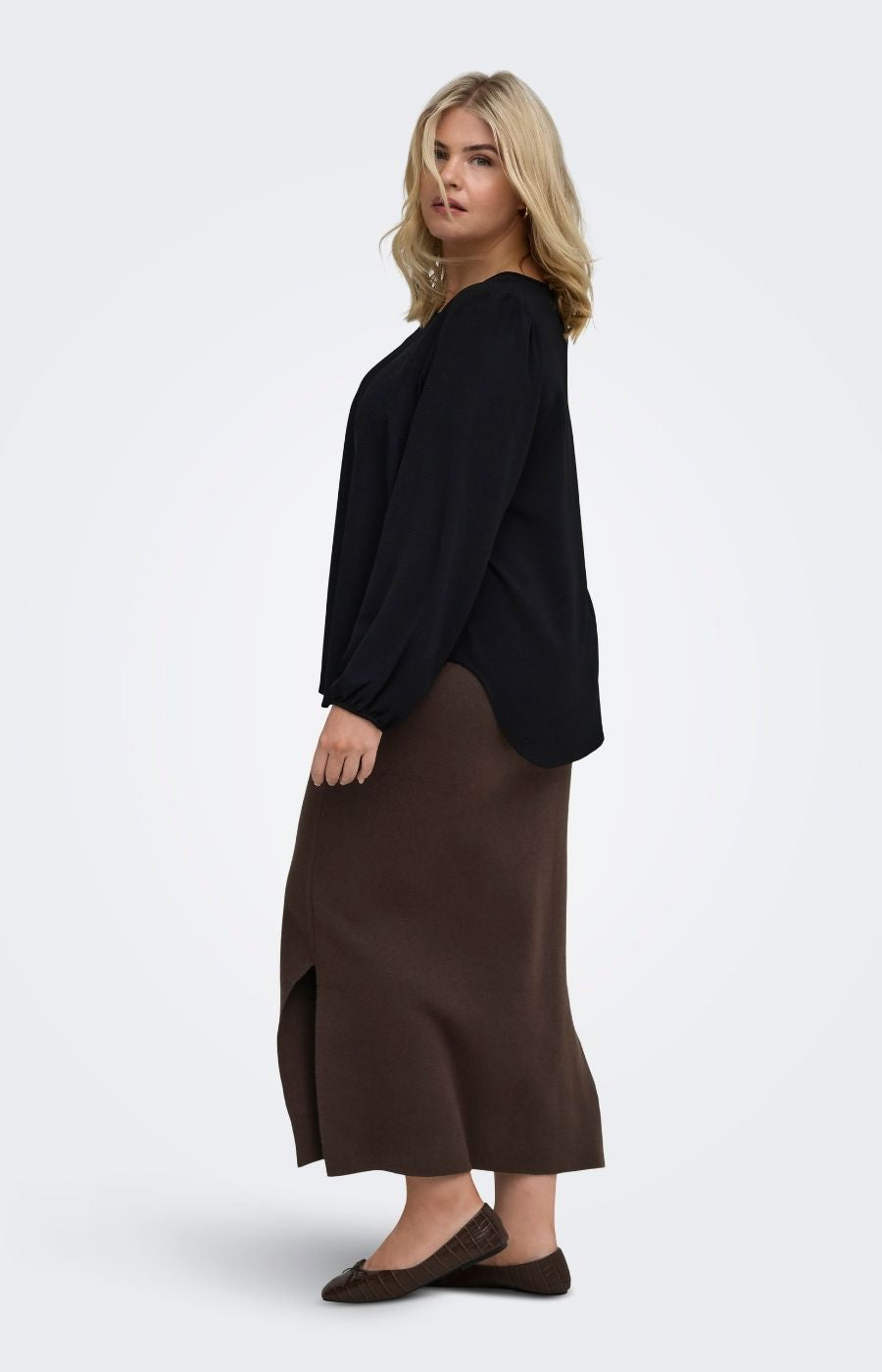 A woman modeling the Only Carmakoma Ribi Skirt in Brown, a brown midi skirt and a black long-sleeve top shown from a side-back angle against a light gray studio background.