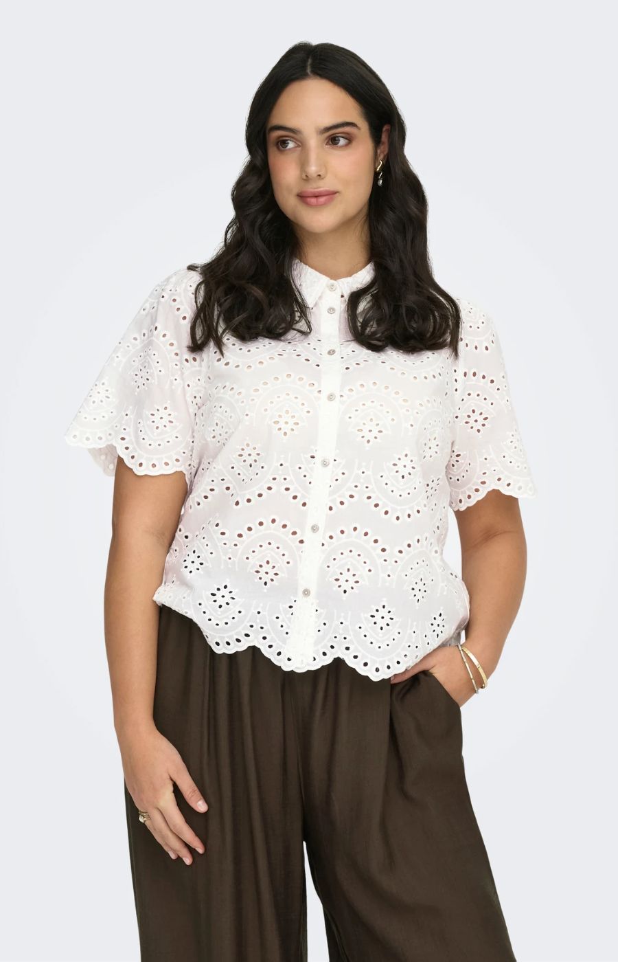 Woman wearing a white lace blouse and brown pants on a light gray background