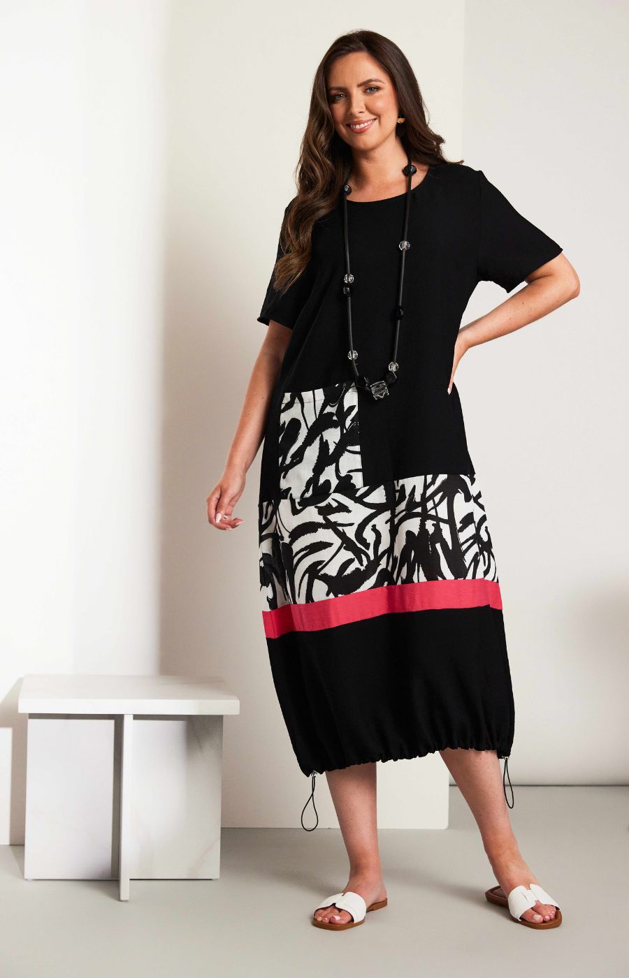 Woman wearing the Ora Dress in Black with Necklace, a black dress with a white, black and pink design on the fron, front pocket, short sleeves and a round neckline against a white background