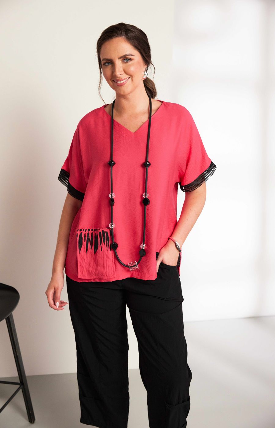 Woman wearing the Ora Feather Print Top in Pink, a pink blouse with black trim, front pocket, V neckline and short sleeves and black trousers against a white background
