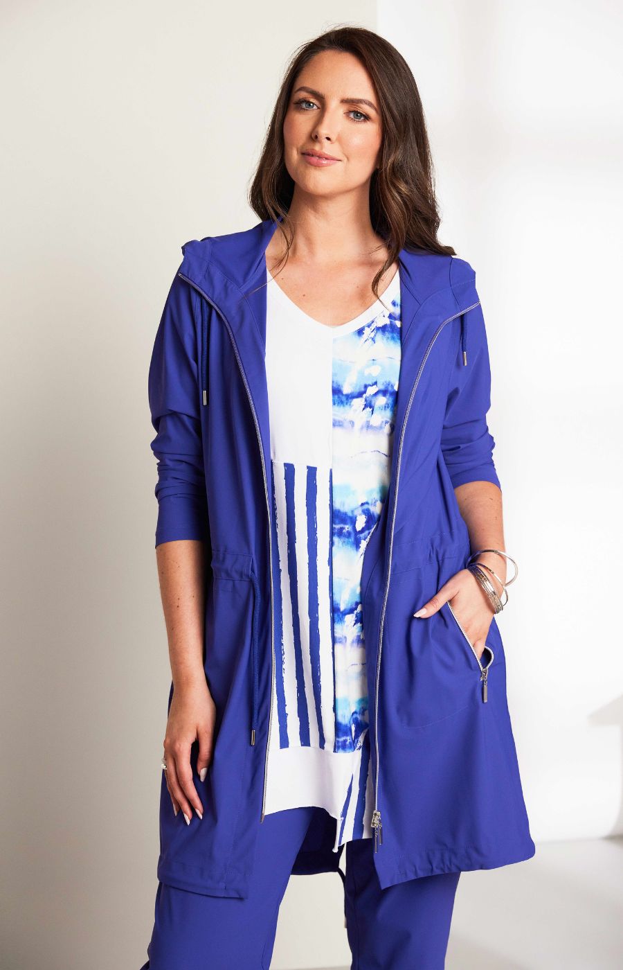 Woman wearing the Ora Hooded Jacket in Blue, with the blue Ora travel trousers a blue outfit with a white background