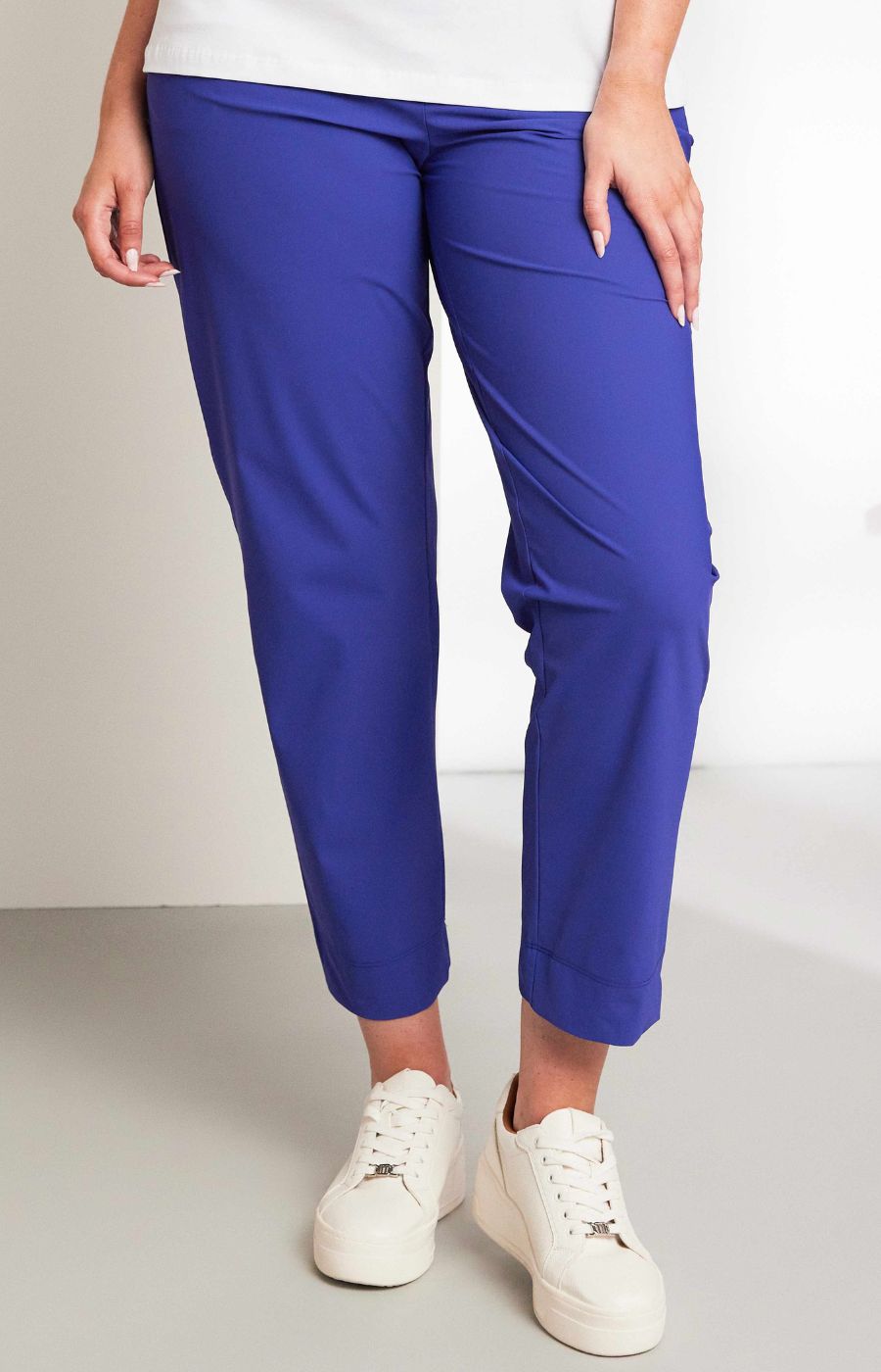 Women wearing the Ora Travel Trousers in Blue, a blue pull up style, full length trouser with an elasticated waistband and side pockets, worn with white sneakers on a plain background