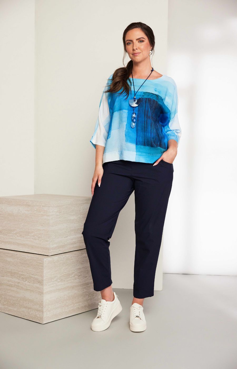 Women wearing the Ora Travel Trousers in Navy, a full length navy trouser with a straight leg, elasticated waistband and side pockets worn with white sneakers and a blue top against a neutral background