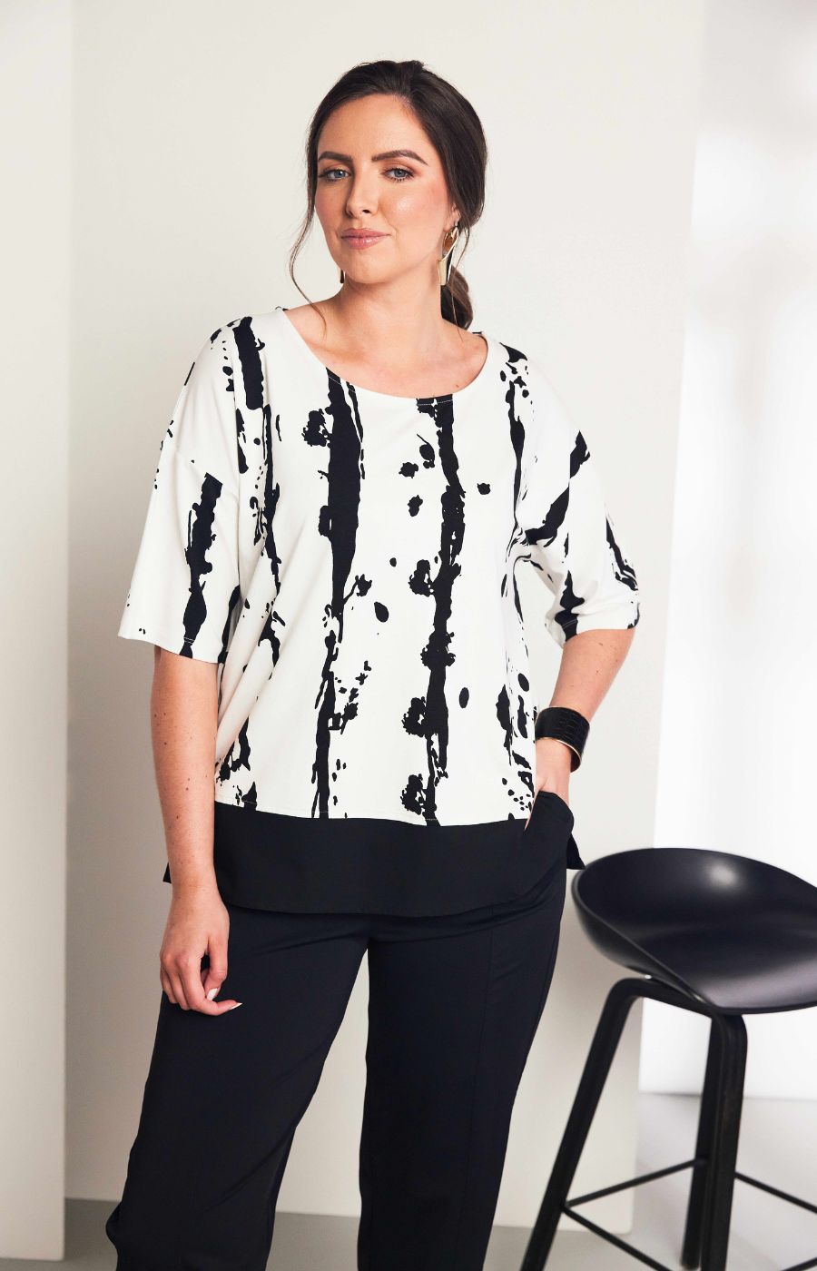 Woman wearing the Ora White and Black Stripe Top, a black and white messy stripe patterned top with 1/2 length sleeves, round neckline, relaxed fit and contrasting hem worn with black trousers.
