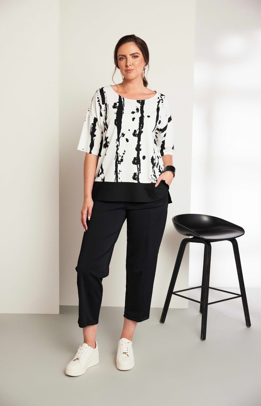Woman wearing the Ora White and Black Stripe Top, a black and white messy stripe patterned top with 1/2 length sleeves, round neckline, relaxed fit and contrasting hem worn with black trousers.