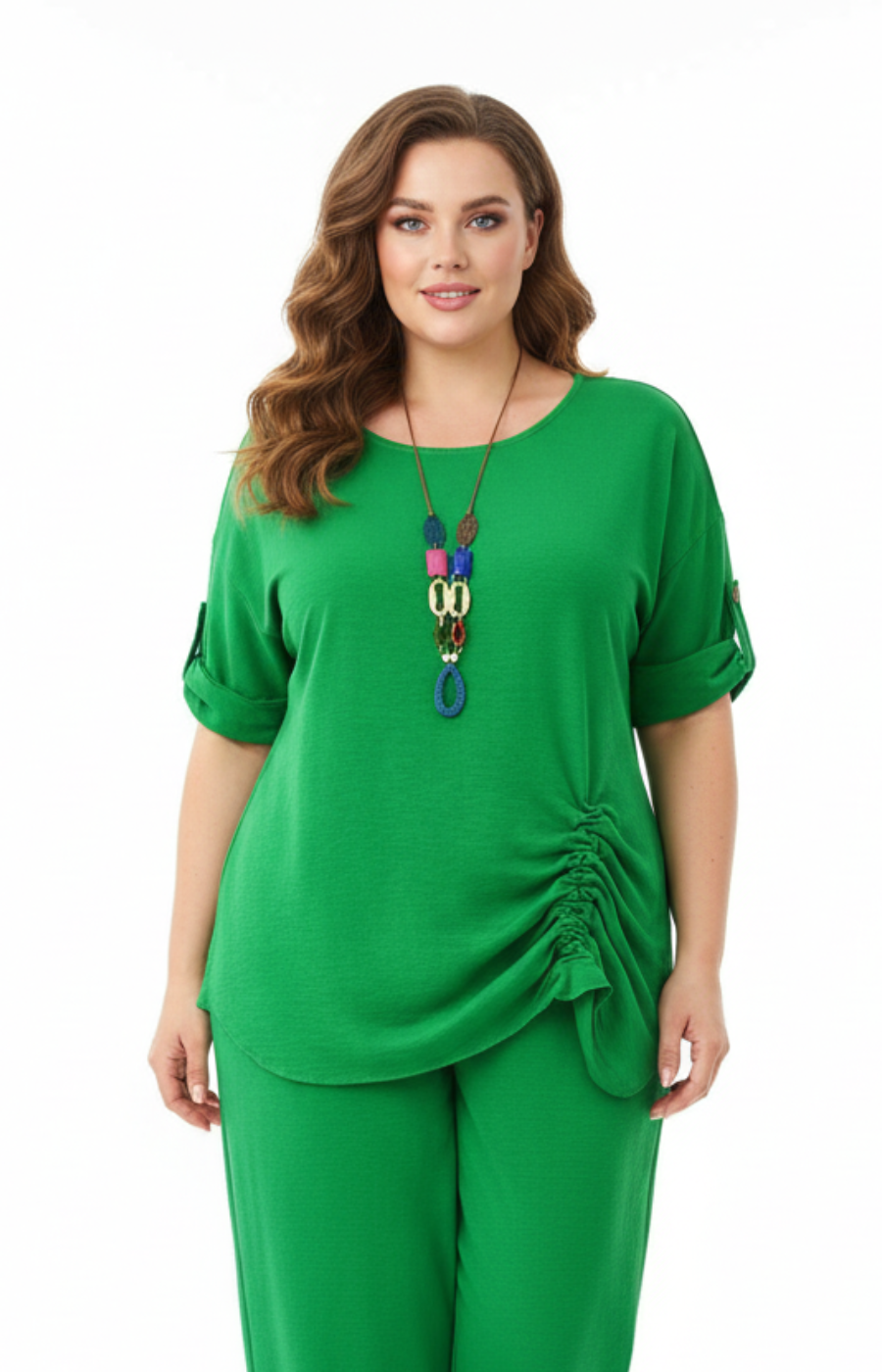 A woman standing against a plain white background wearing a green blouse with rolled-up sleeves and a gathered detail on one side, paired with matching green cropped pants. Accessorized with a long, colorful pendant necklace.