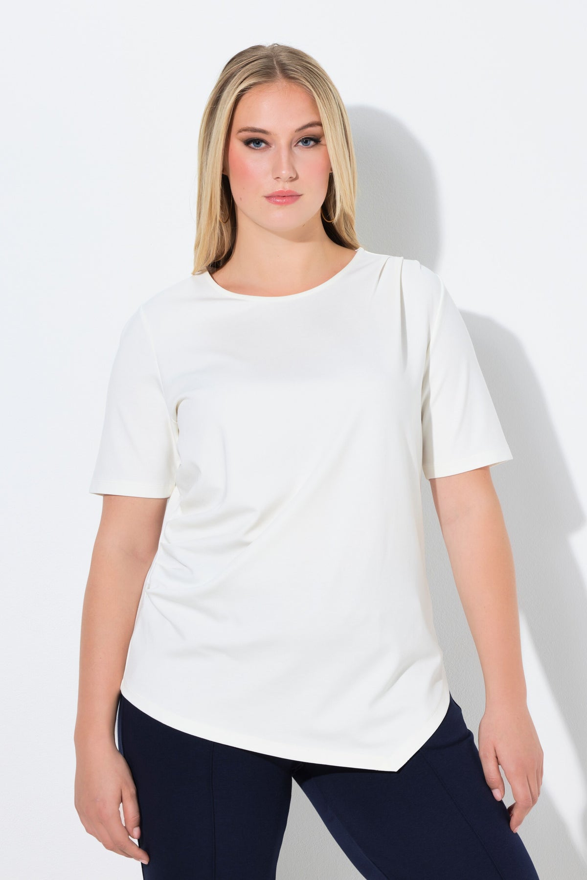 Woman wearing the Ulla Popken Pointed Hem T-Shirt in Off White, a white t-shirt with a round neckline, a pointed hem and draping on the shoulder and side worn with dark pants on a white background