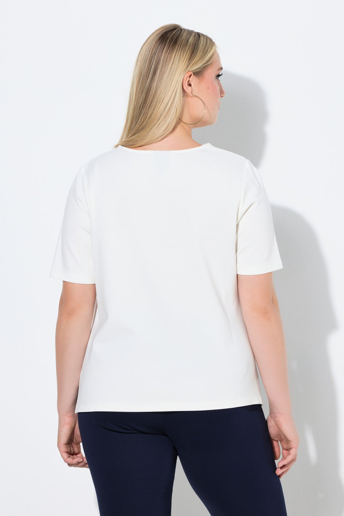Woman wearing the Ulla Popken Pointed Hem T-Shirt in Off White, a white t-shirt with a round neckline, a pointed hem and draping on the shoulder and side worn with dark pants on a white background