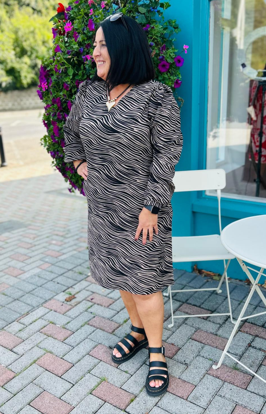 Woman in a patterned dress standing outdoors with a blue building and white chairs in the background.