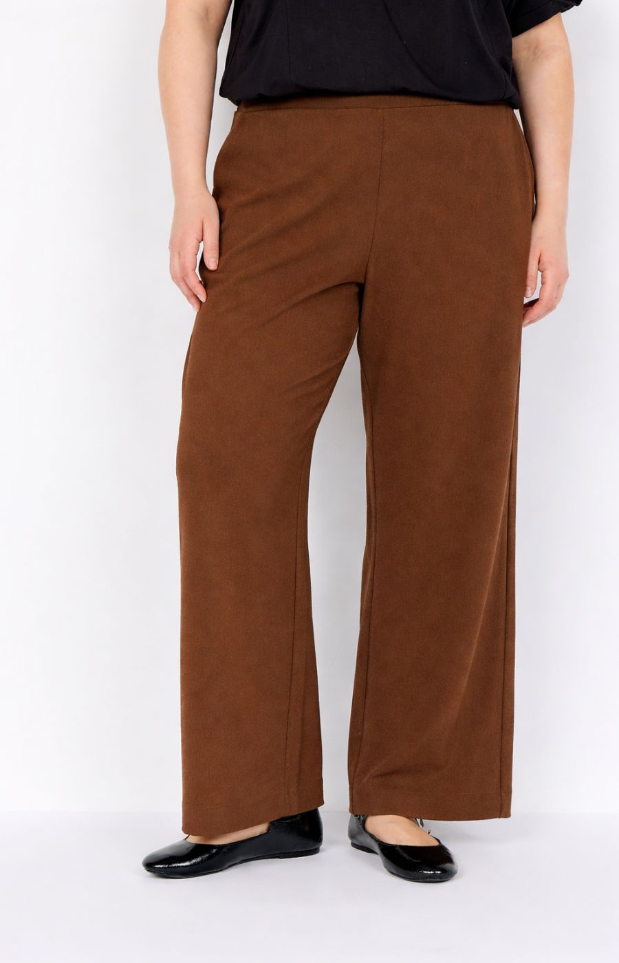 Person wearing Wasabi Kufa Trousers in Brown, a straight, full length trouser with side pockets and black shoes on a white background