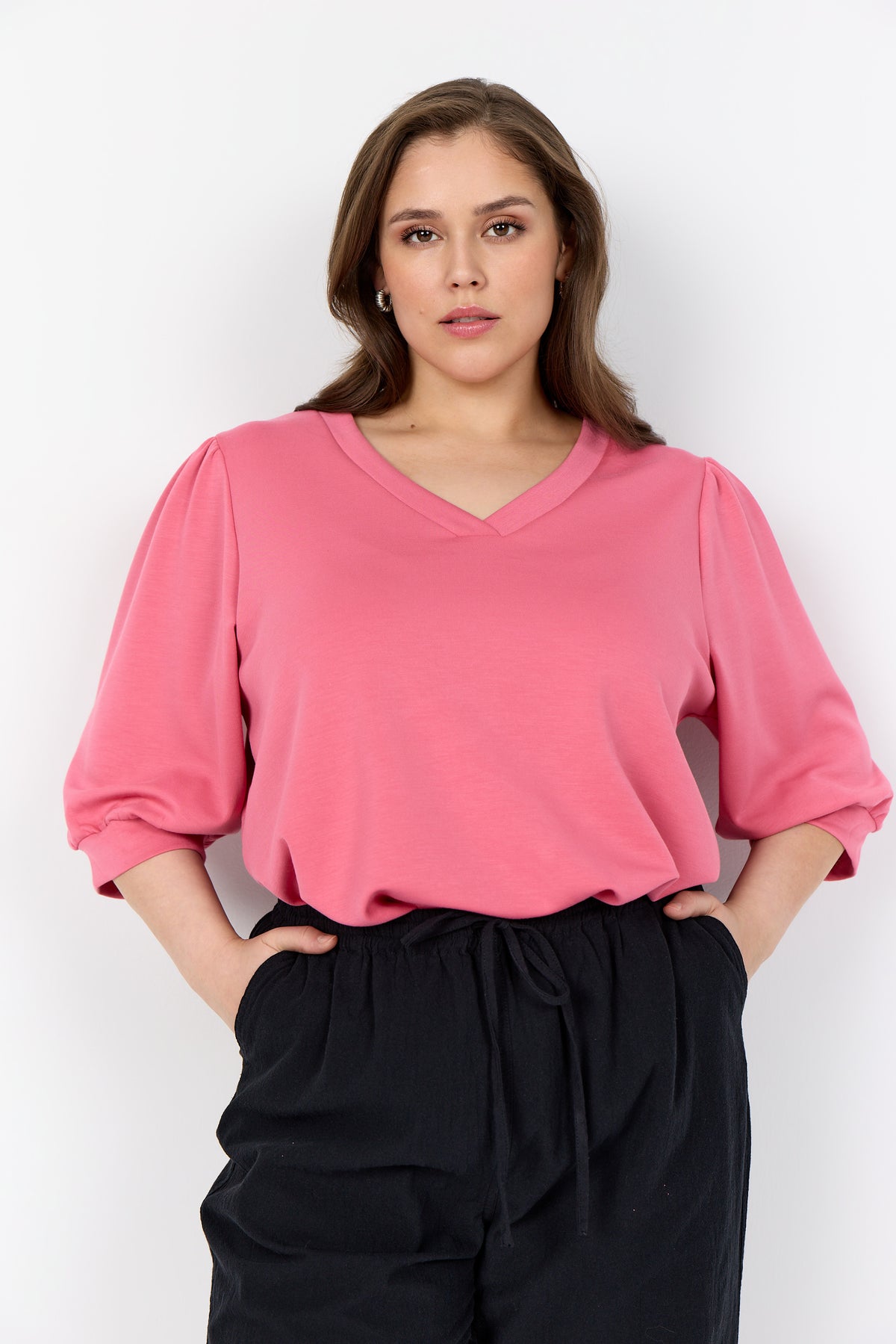 Woman wearing a Wasabi Sabina Blouse in Pink, a V neck top with 1/2 length puff sleeves with a cuff and black pants on a white background