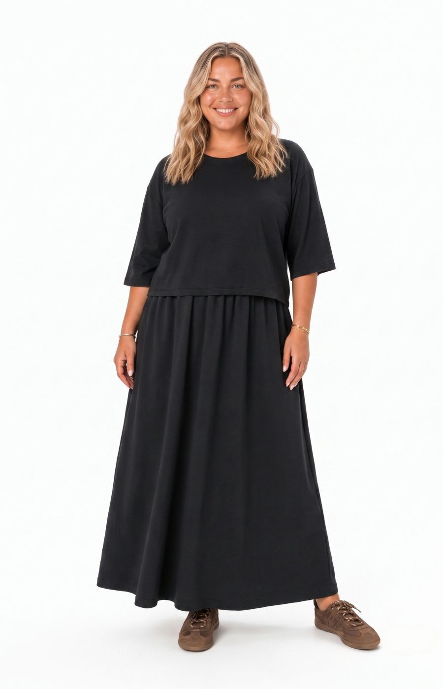 A woman modeling the Wasabi Sabina Dress in Black, a black two-piece look outfit consisting of a short-sleeve crewneck top and a matching long pleated skirt, paired with brown shoes, against a plain white background. This image has been created using AI.