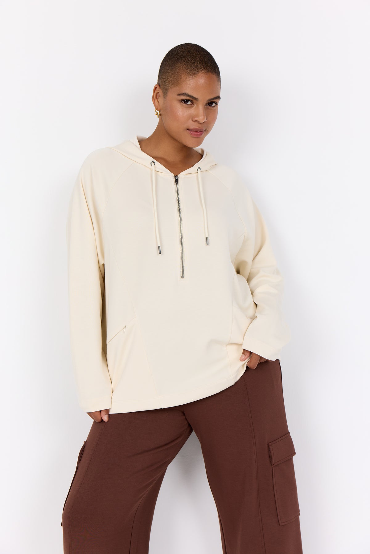 Person wearing a Wasabi Sabina Hoodie in Cream, a 1/2 zip hoodie with front zipped pockets and brown pants against a white background