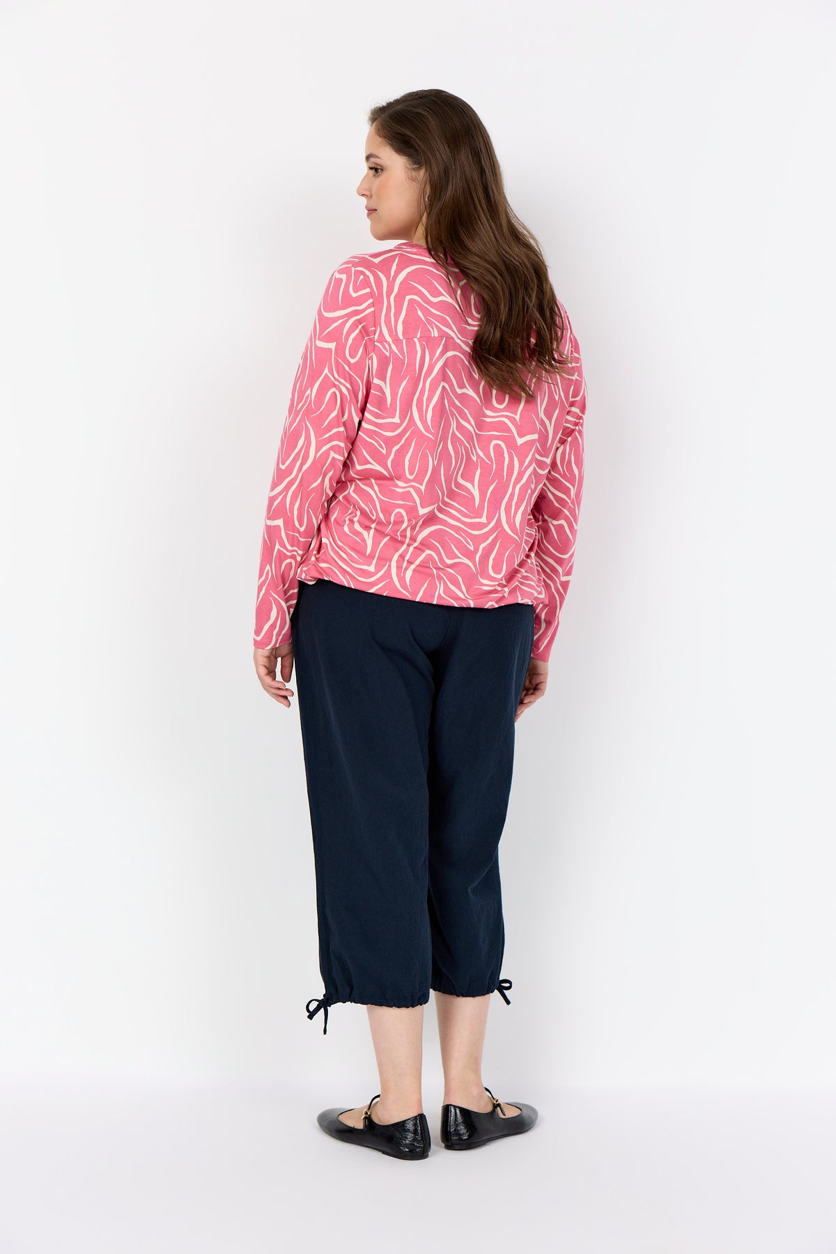 Woman wearing Wasabi Saliha Blouse in Pink, a blouse with long sleeves and an elasticated hem and navy pants on a white background