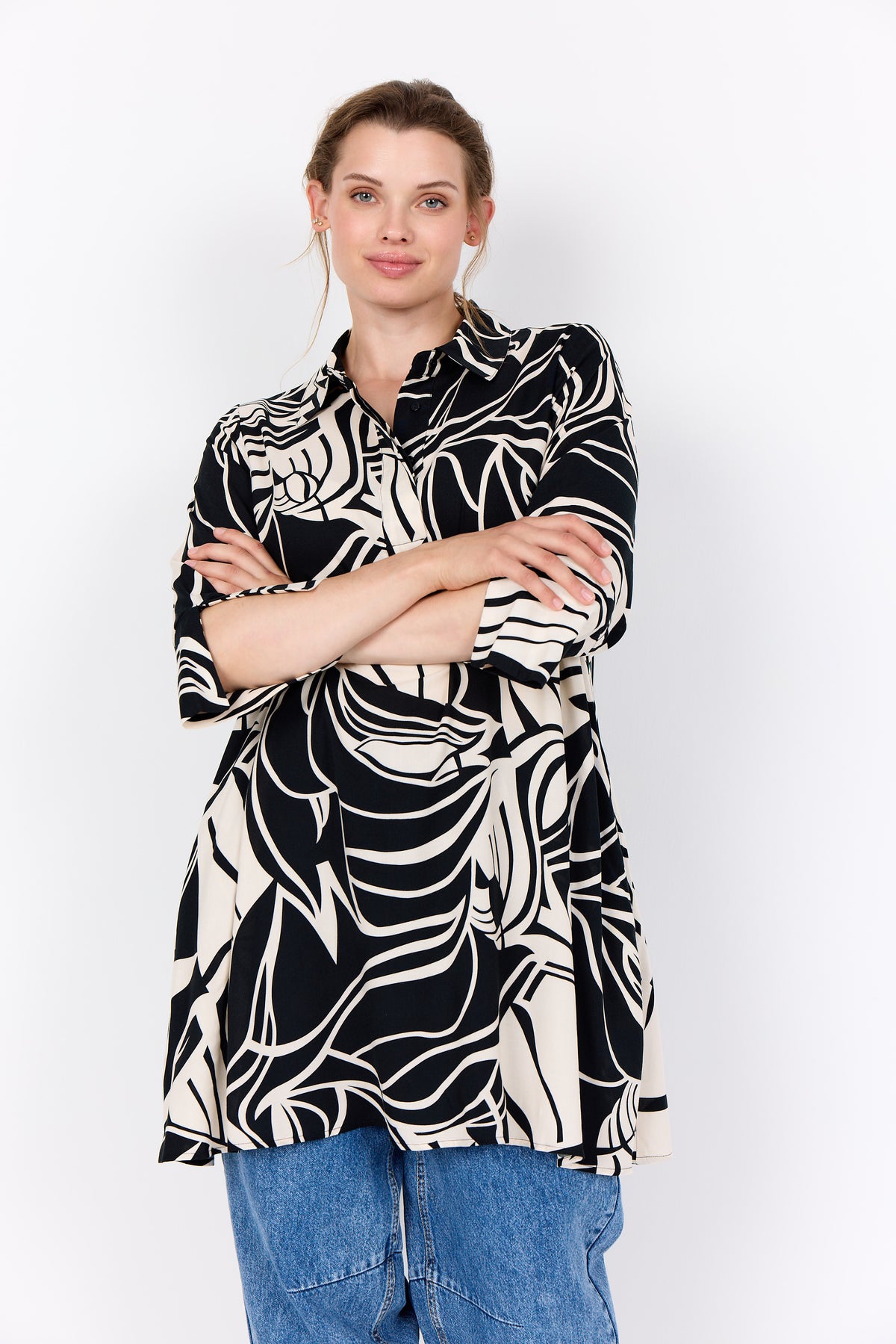 Woman wearing Wasabi Samena Tunic in Black, an A-line shape tunic with an all over print, 3/4 length sleeves, a short button placket and an all over print ,worn with blue jeans on a white background