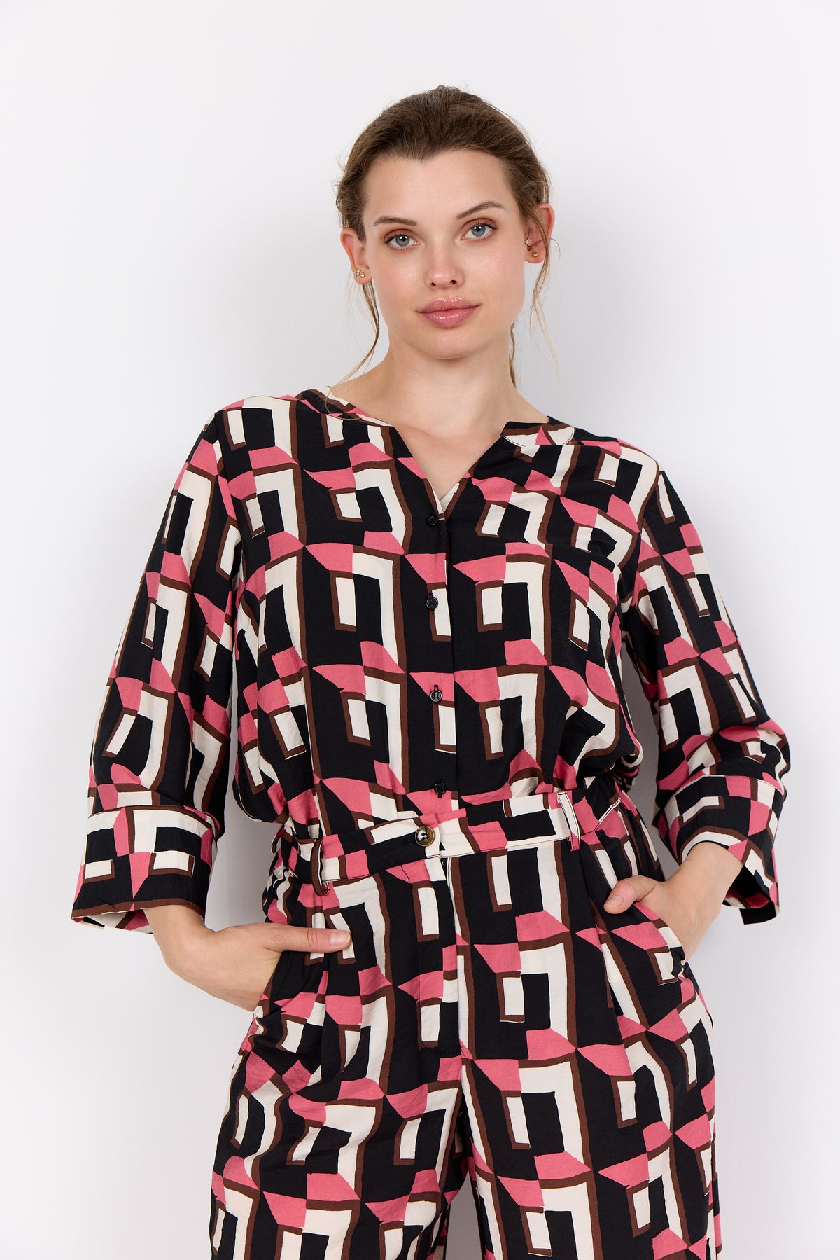 Woman wearing Wasabi Sarena Shirt in Pink with geometric design, V neckline, button down front and 3/4 length sleeves with the matching trousers on a white background