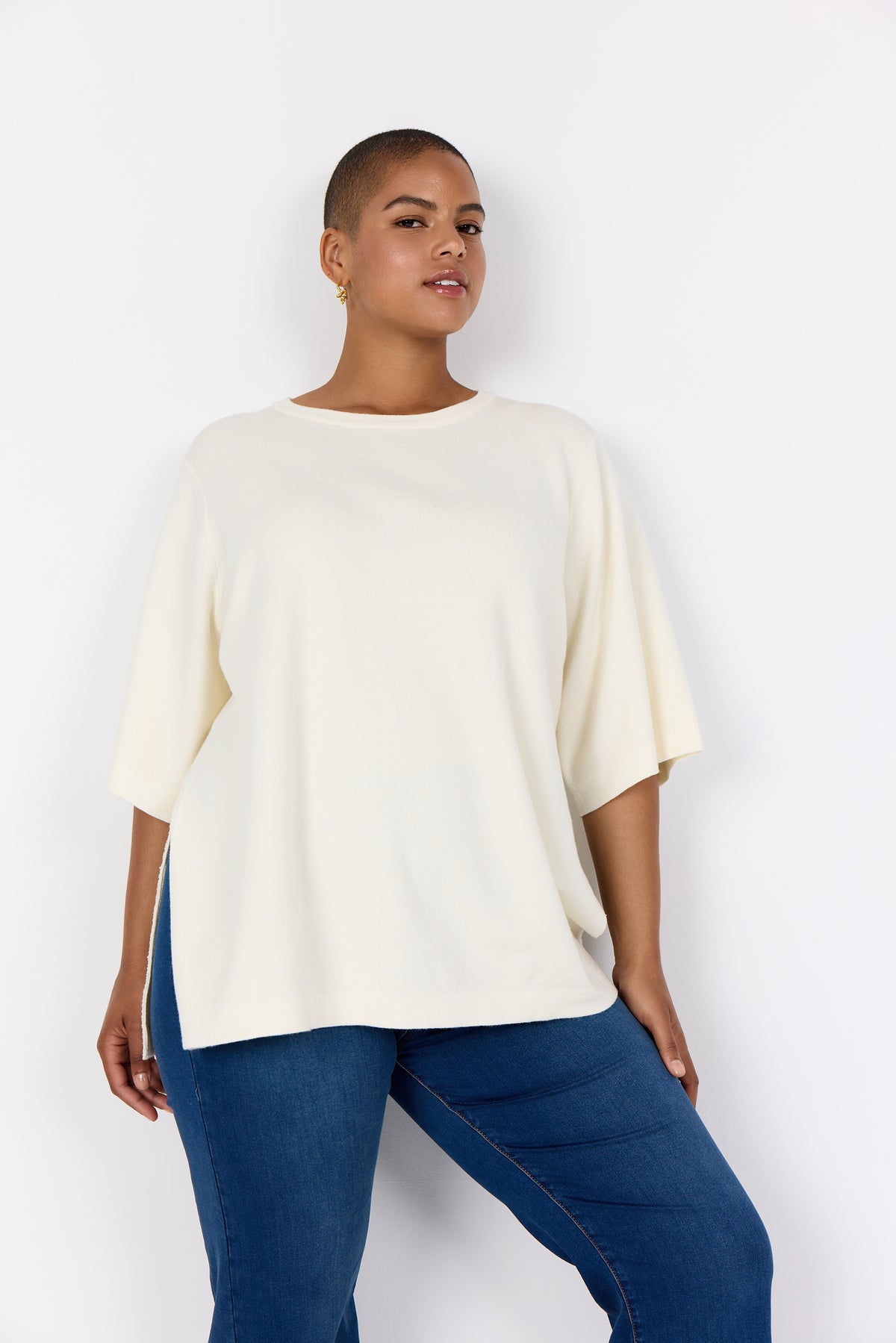 Woman wearing Wasabi Sikka Jumper in Cream, a jumper with 1/2 sleeves, round neckline and side slits with blue jeans against a white background