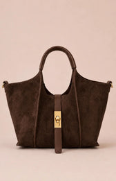 A dark brown suede tote bag with a central gold-tone turn-lock clasp and a matching hanging strap, set against a solid pale pink background.