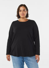 A woman modeling the Zizzi Long Sleeve Top in Black, a black long-sleeve crewneck top with a relaxed fit, paired with blue jeans, against a plain light gray studio background.