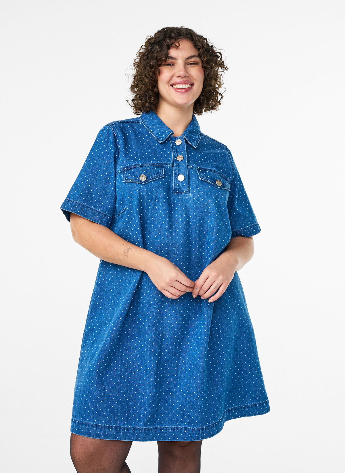 Woman wearing the Zizzi Misha Denim Dress, A blue polka dot dress with a shirt collar, short button placket, knee length and with short sleeves on a white background