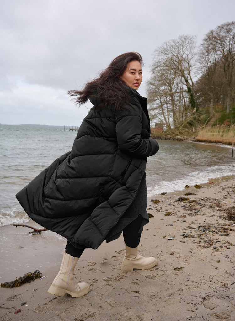 Woman in a black puffer coat standing on a beach with water and trees in the background